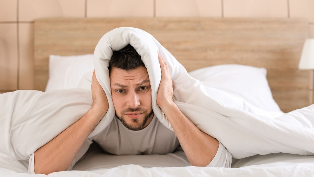 Man laying on his bed, covering his ears with the covers